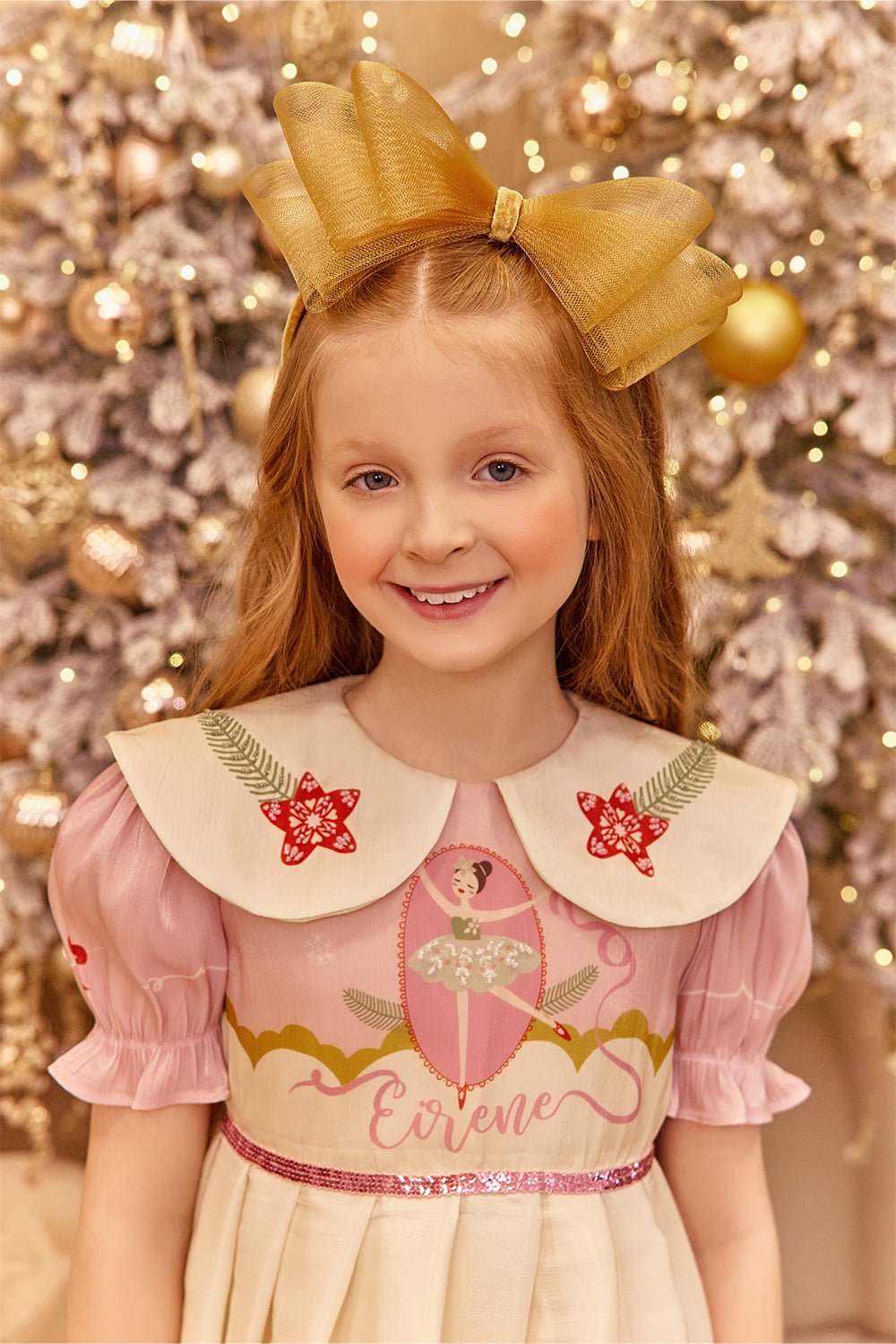 Young girl in a pink dress with a large bow in her hair, standing in front of a decorated Christmas tree.