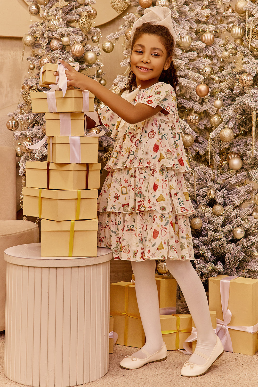 Young girl in a floral dress standing next to a stack of gift boxes with a decorated Christmas tree in the background.