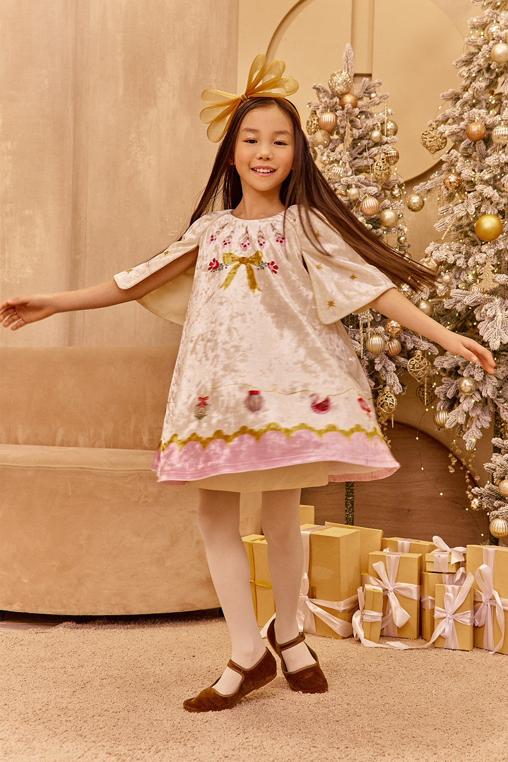 Young girl in a white dress with floral embroidery standing in front of a decorated Christmas tree.