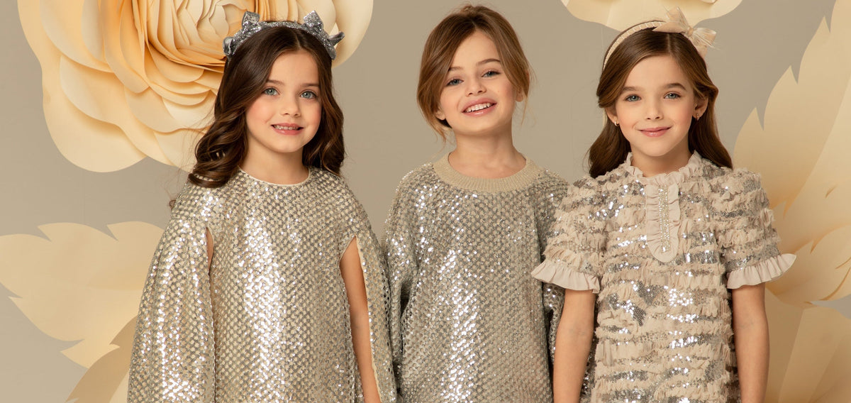 Three young girls modeling sequined dresses and tops against a beige floral backdrop. The girl on the left wears a silver sequined tunic dress with a silver bow headband.
