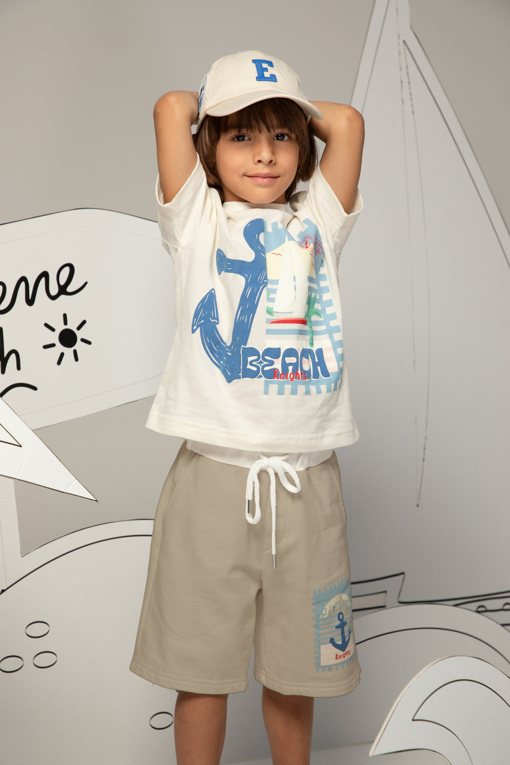 Child wearing a white t-shirt with a blue anchor design and beige shorts, standing against a gray background.