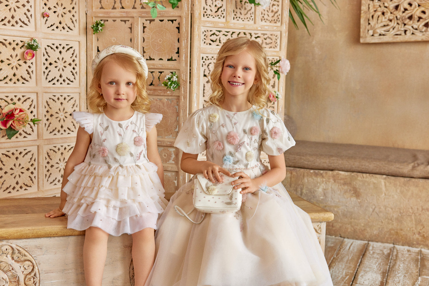 Two young girls in white dresses sitting on a wooden bench with a decorative wall behind them.