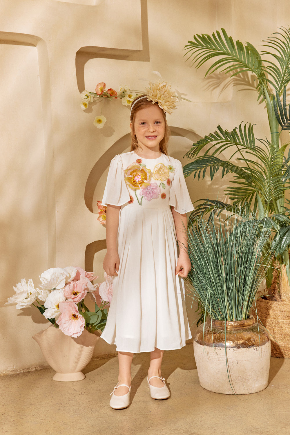 Young girl in a white dress with floral design standing in a decorative setting with plants and flowers.