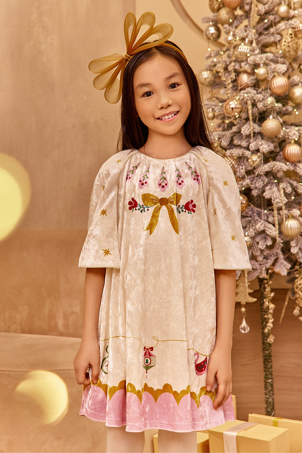 A girl wearing an ivory and pink velvet Christmas dress with hand-embellishments, standing near a Christmas tree.