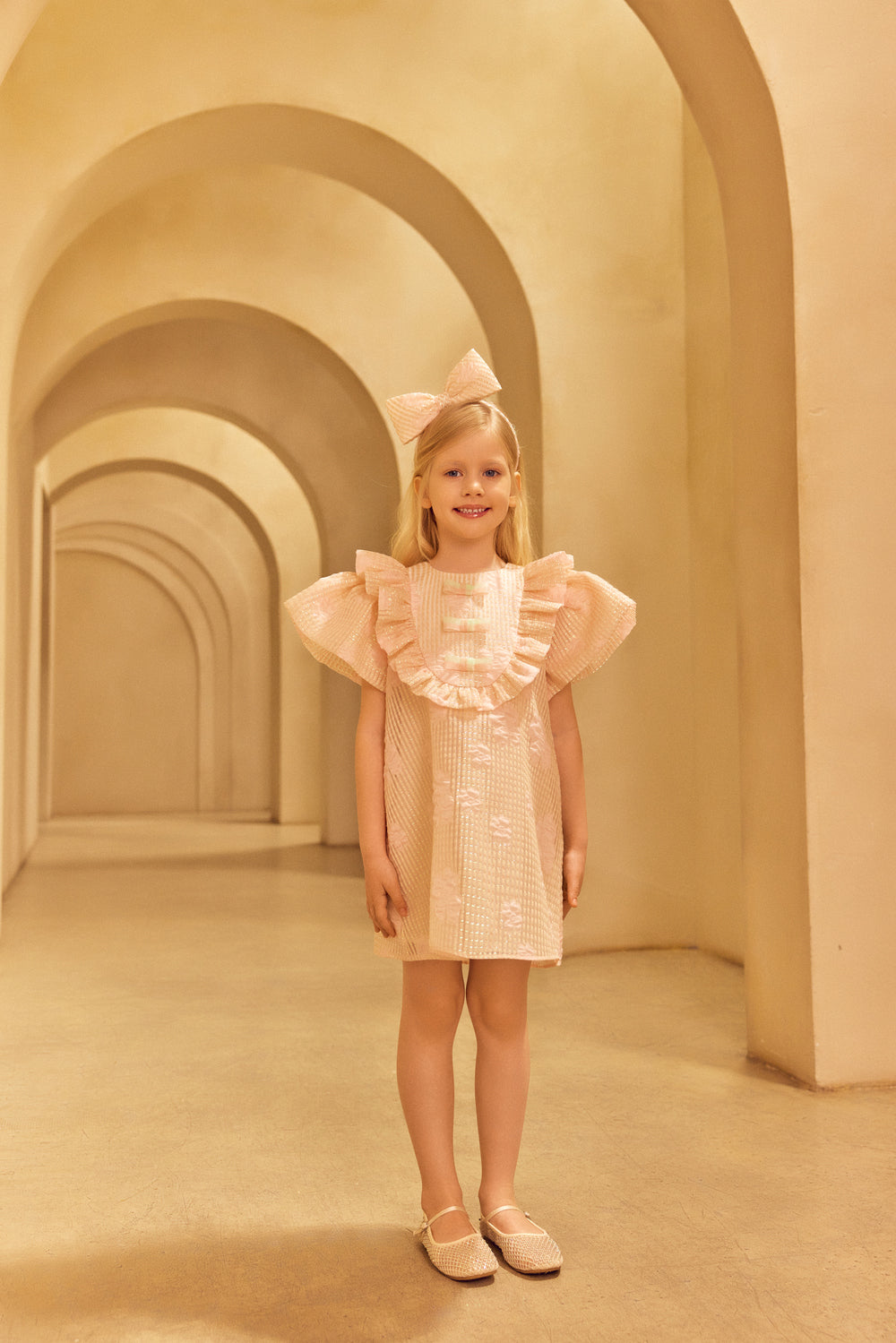 Young girl in a pink dress and a pink headband standing in a beige archway corridor