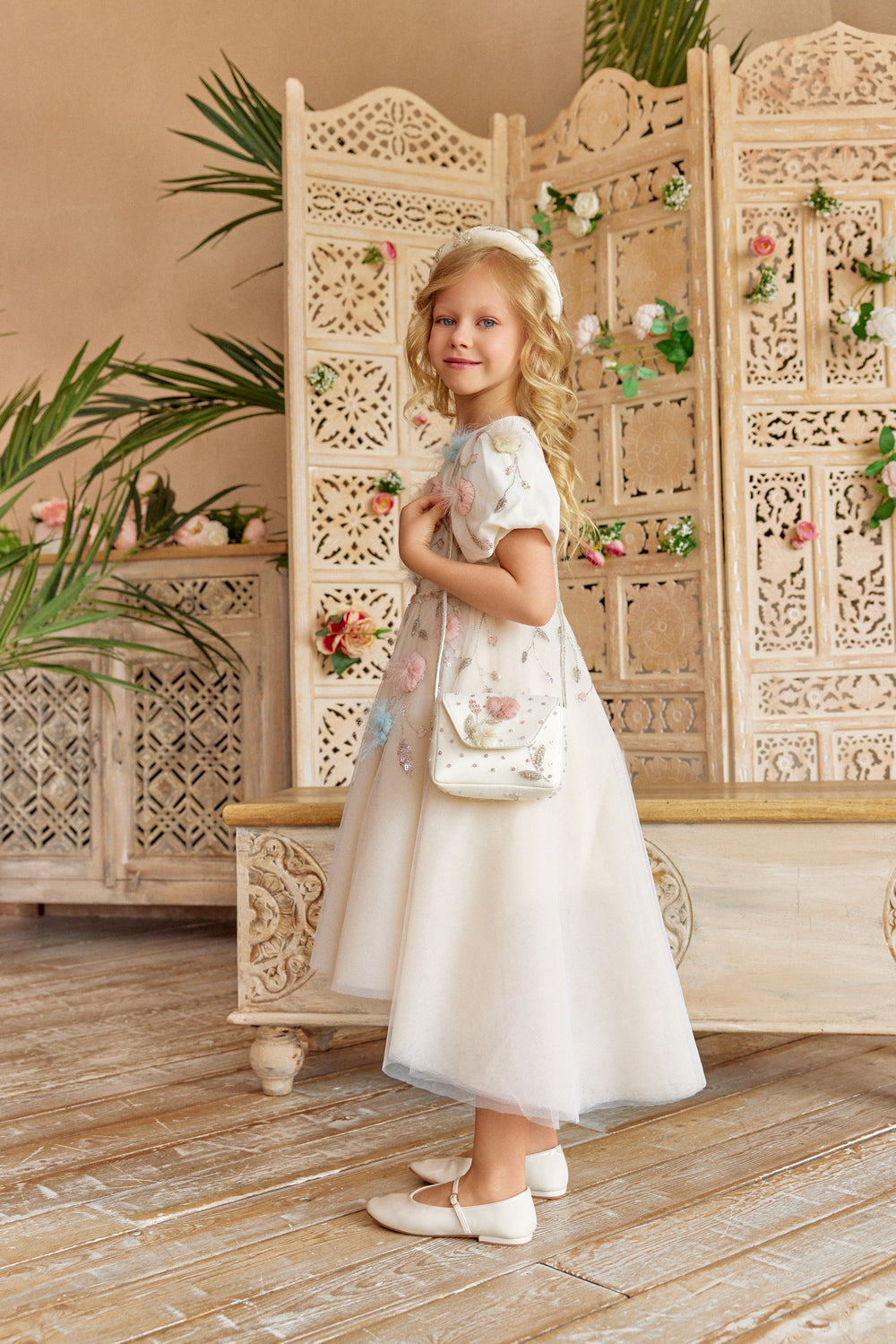 Young girl in a floral dress with a decorative bag, sitting on a wooden bench.