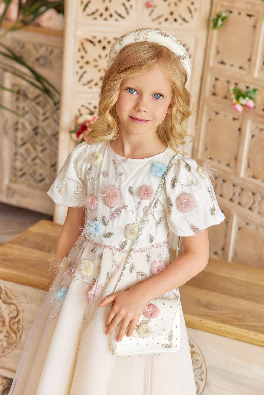 Young girl in a floral dress with a decorative headband, sitting on a wooden bench.