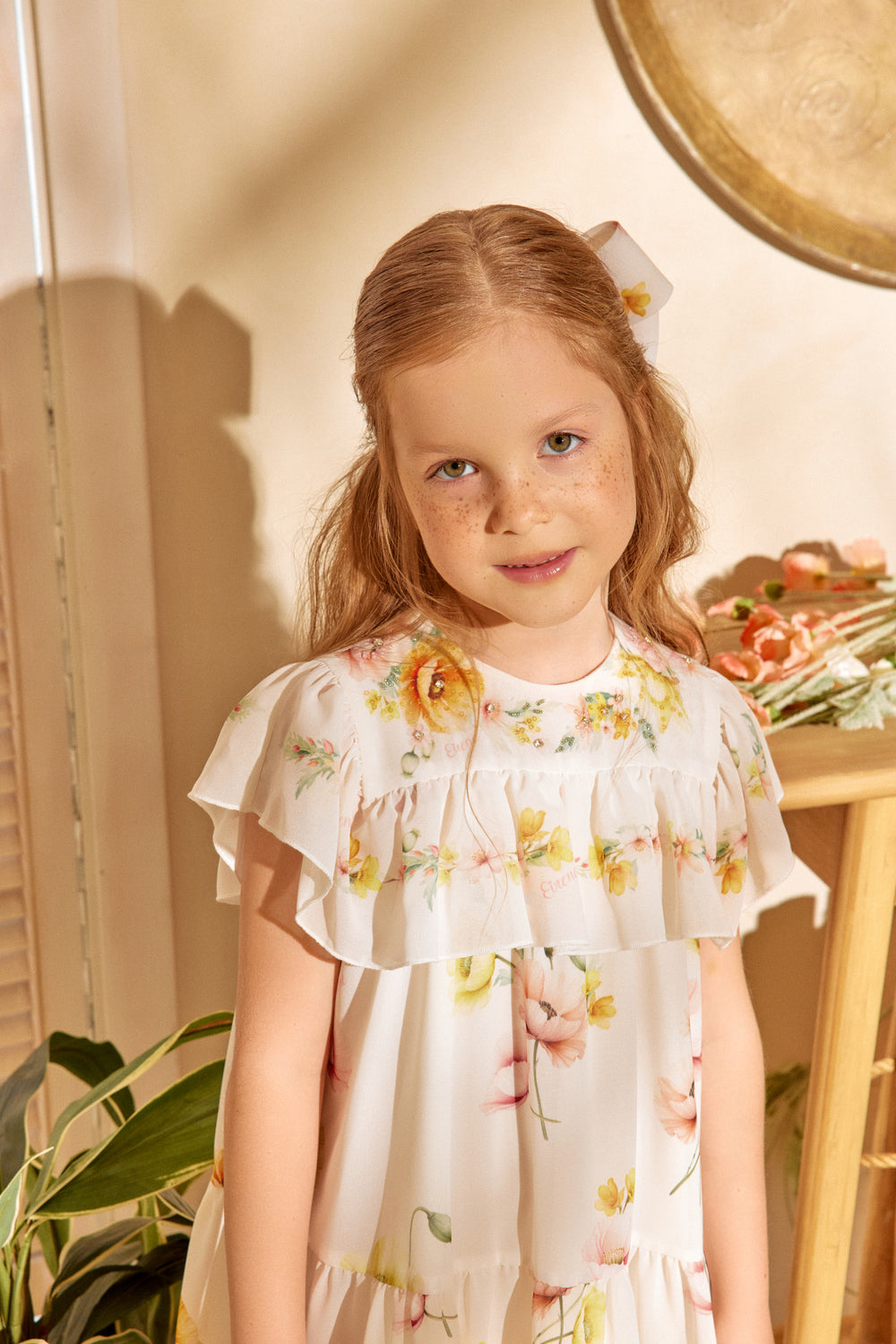 Young girl wearing a floral dress standing in a softly lit room with plants.