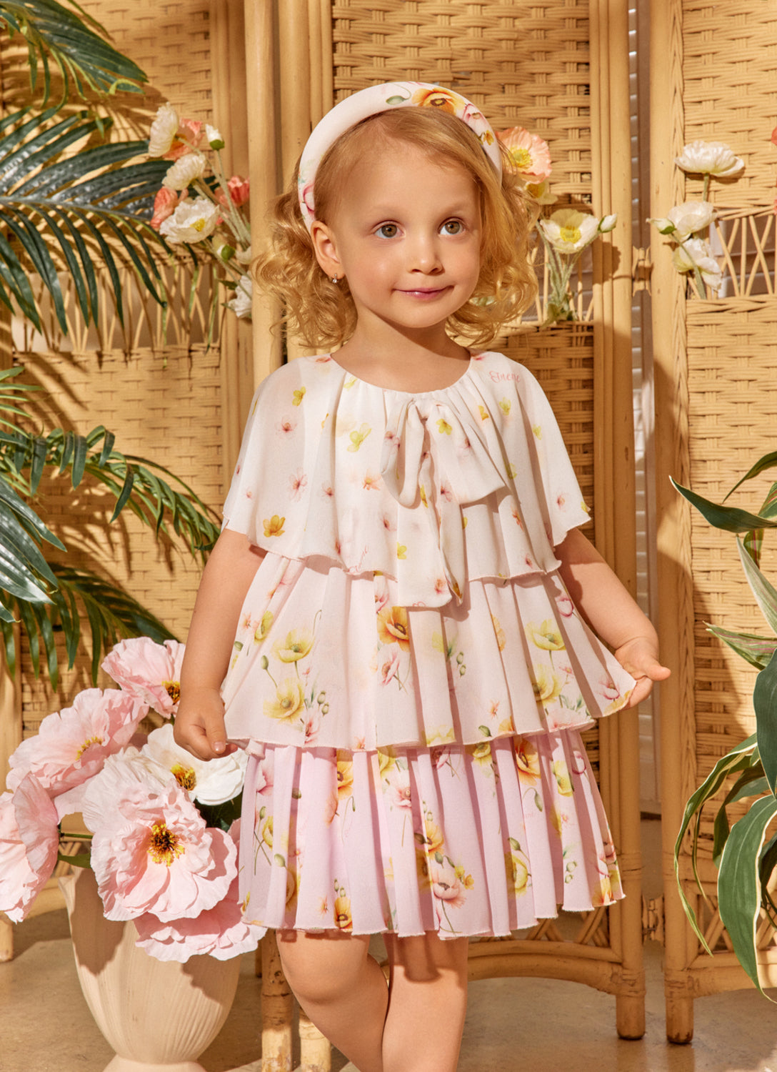 Young girl in a floral dress standing in a decorative setting with plants and flowers.