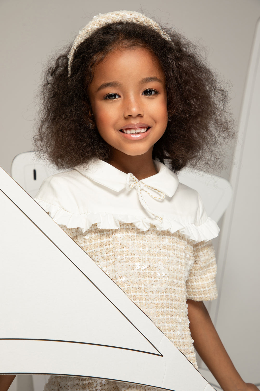 Young girl in a white dress with a large bow sitting on a white chair.