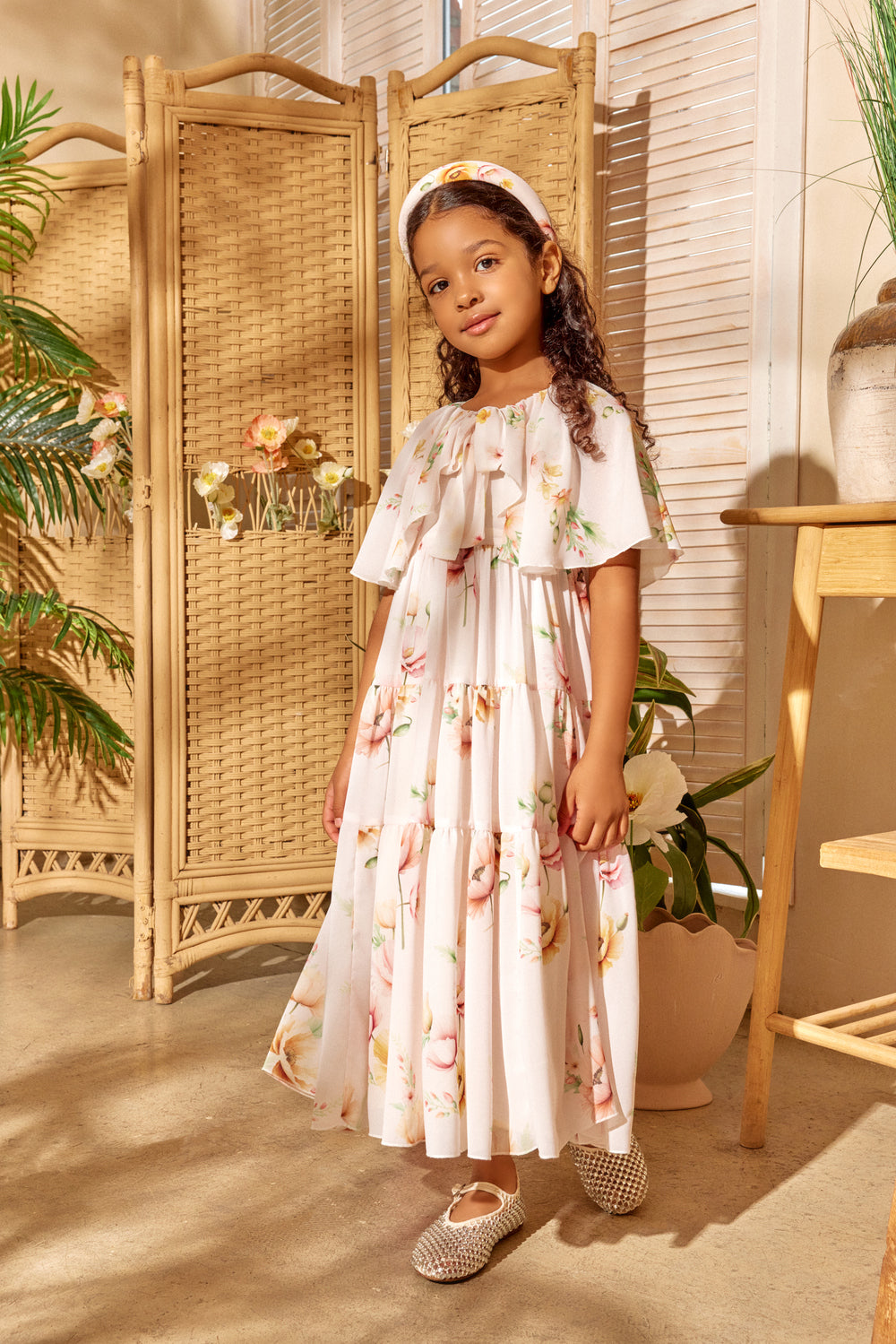 Young girl in a floral dress standing in a room with plants and a woven screen.