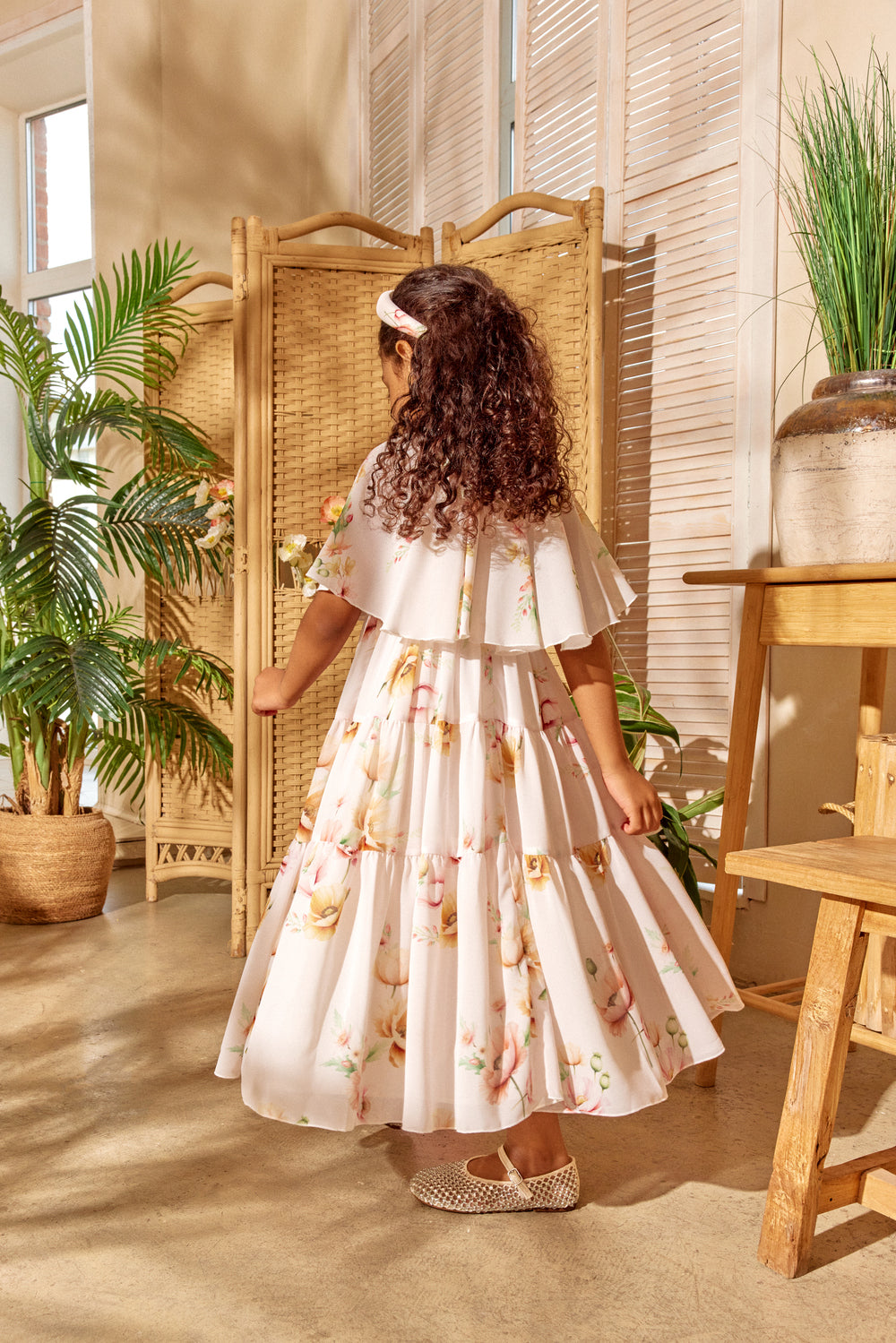 Girl in a floral dress standing in a sunlit room with plants and wooden furniture.