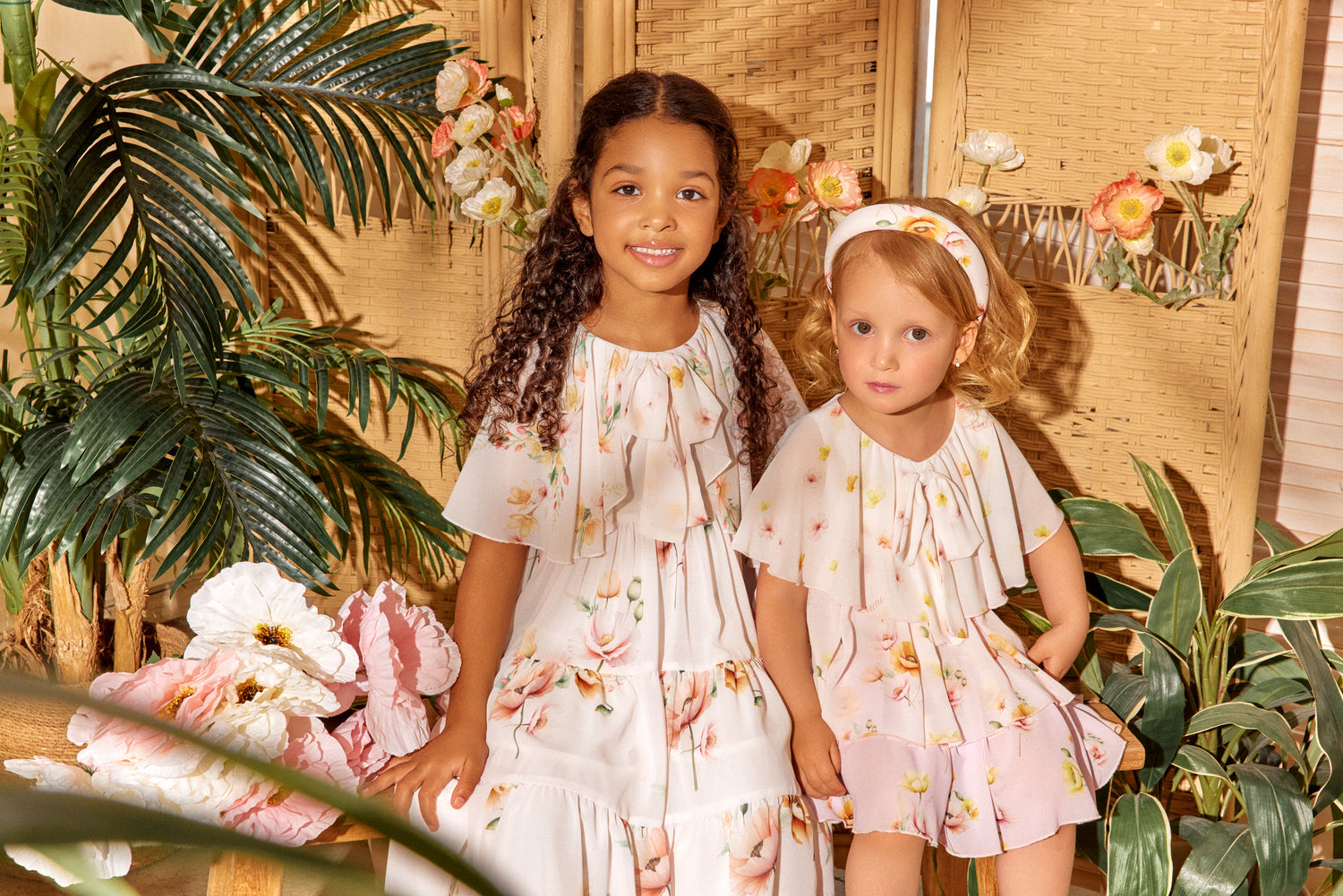 Two young girls in floral dresses sitting together with plants and flowers in the background