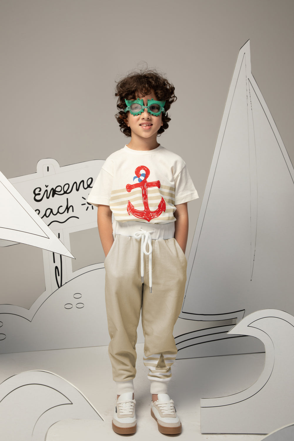 Child wearing a white t-shirt with a red anchor design and beige pants, standing against a gray background.