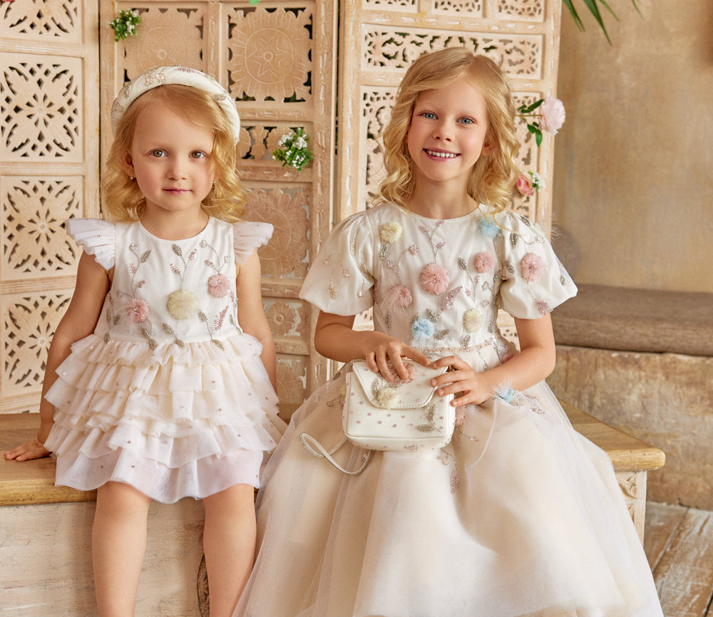 Two young girls in ivory dresses with floral details sitting on a wooden bench.