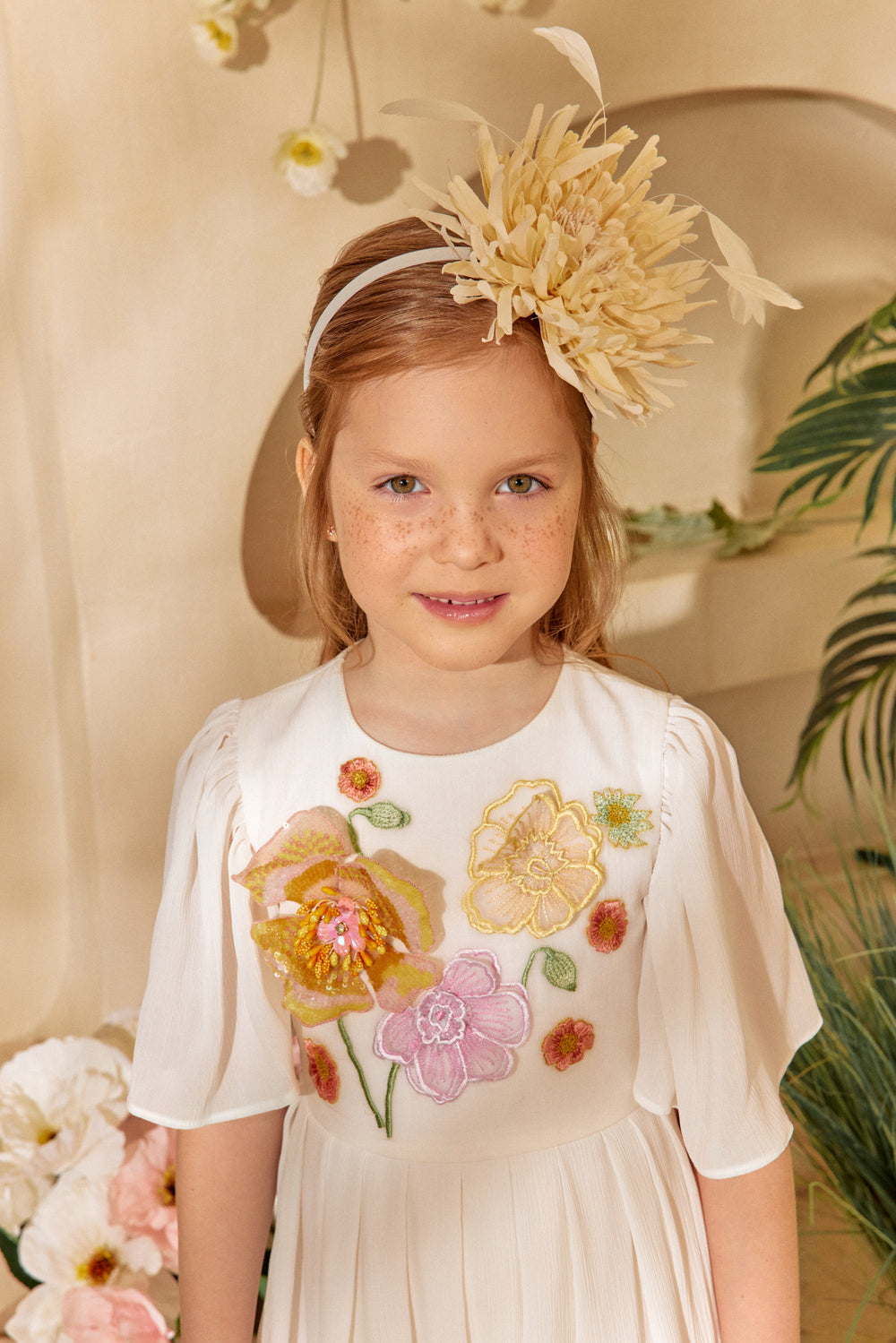 Young girl wearing a floral dress and headband with flowers, standing against a neutral background.