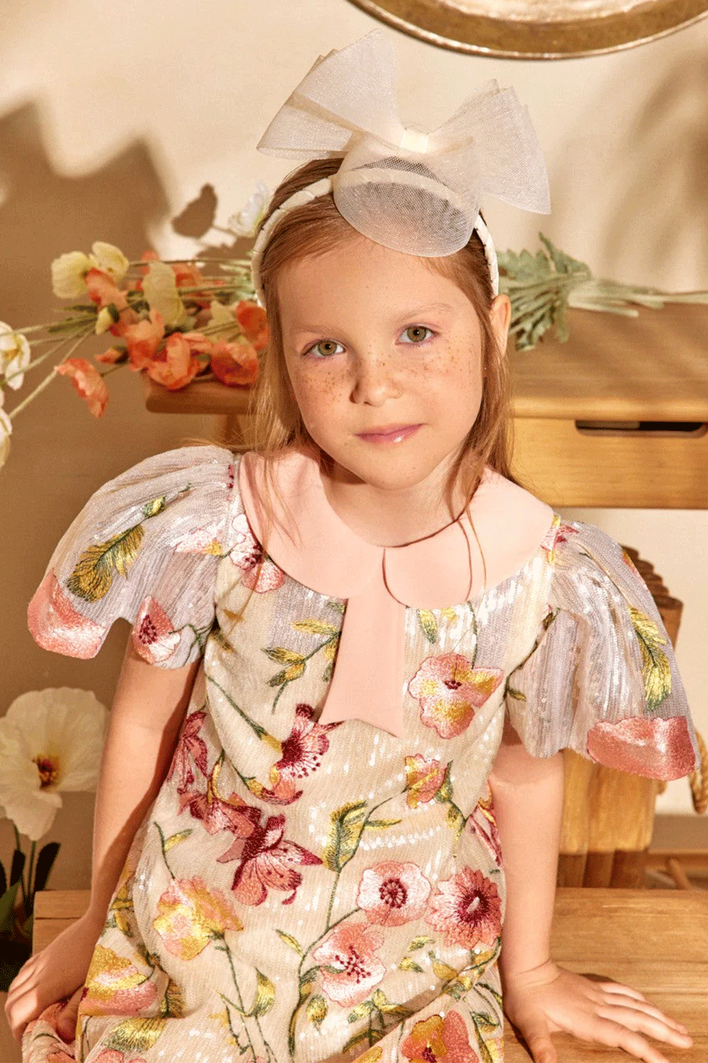 فستان عاجي اللون مزين بالترتر ومطرز بالزهور، وأكمام معدنية مطوية، وياقة بيتر بان وردية ناعمة مزينة بفيونكة.