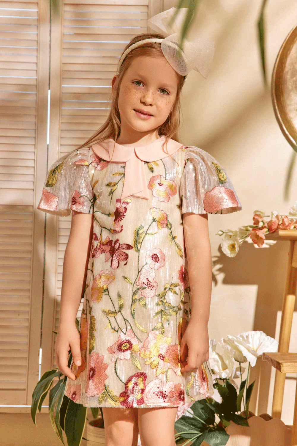 فستان عاجي اللون مزين بالترتر ومطرز بالزهور، وأكمام معدنية مطوية، وياقة بيتر بان وردية ناعمة مزينة بفيونكة.